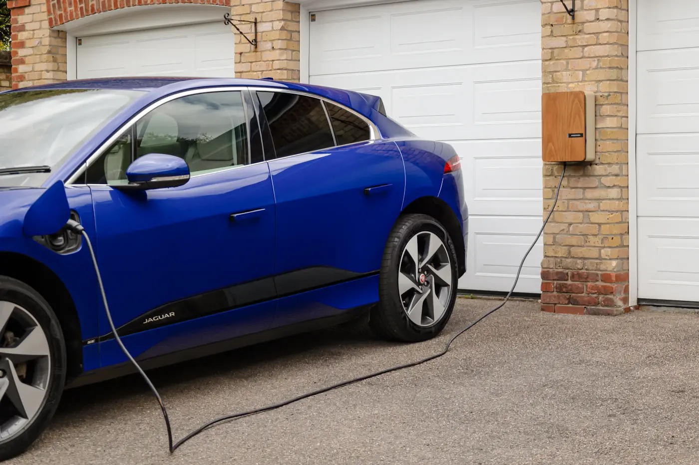 Une voiture électrique bleue branchée devant le garage d'une maison, à côté d'une borne murale.